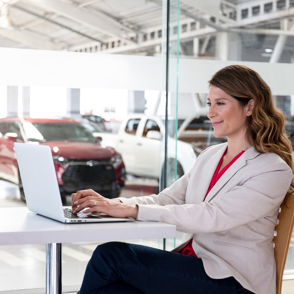 Mujer sonriendo mientras solicita una cotización en línea desde una laptop en un concesionario Chevrolet, con vehículos de fondo.