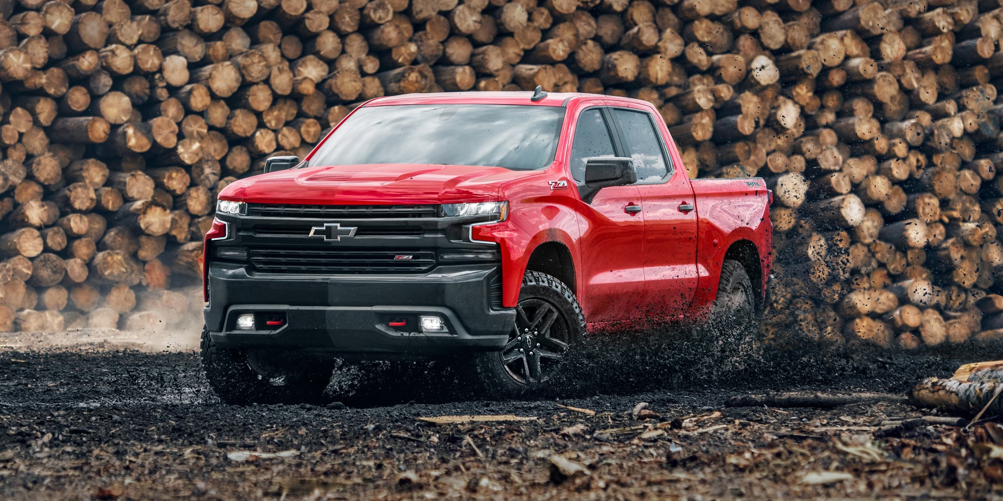 Chevrolet Silverado color Rojo Brillante atravesando un camino de tierra con troncos apilados de fondo.