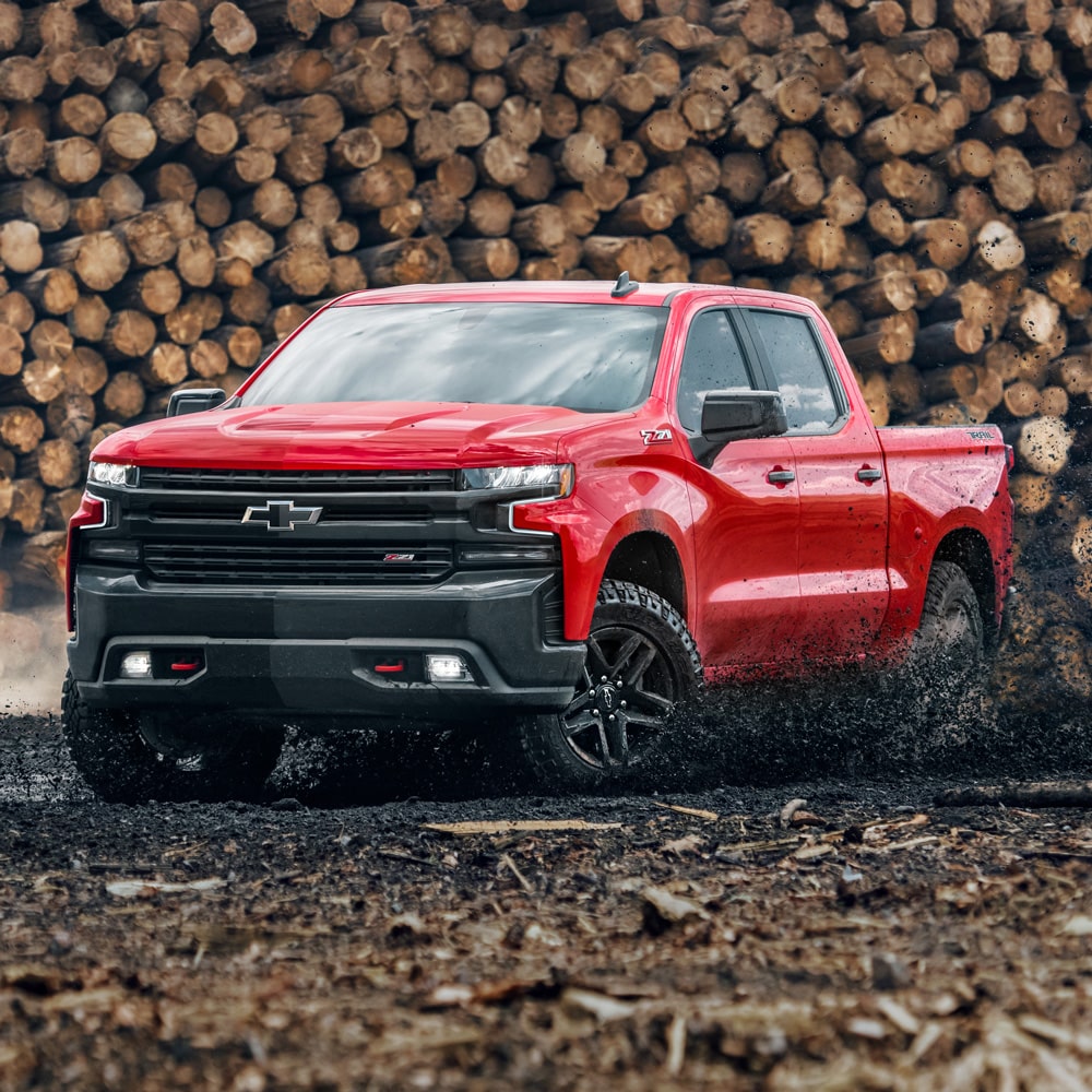 Chevrolet Silverado color Rojo Brillante atravesando un camino de tierra con troncos apilados de fondo.