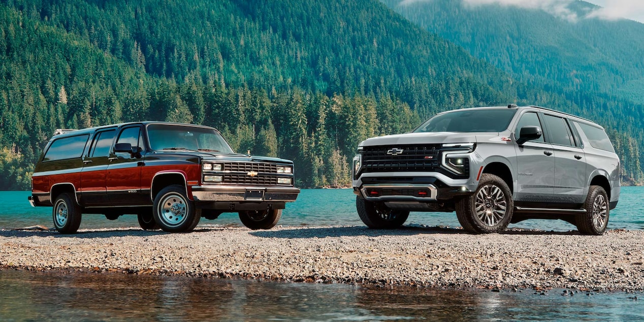 Chevrolet Suburban clásica junto a la nueva Suburban Z71, exhibidas frente a un lago rodeado de montañas y bosques.